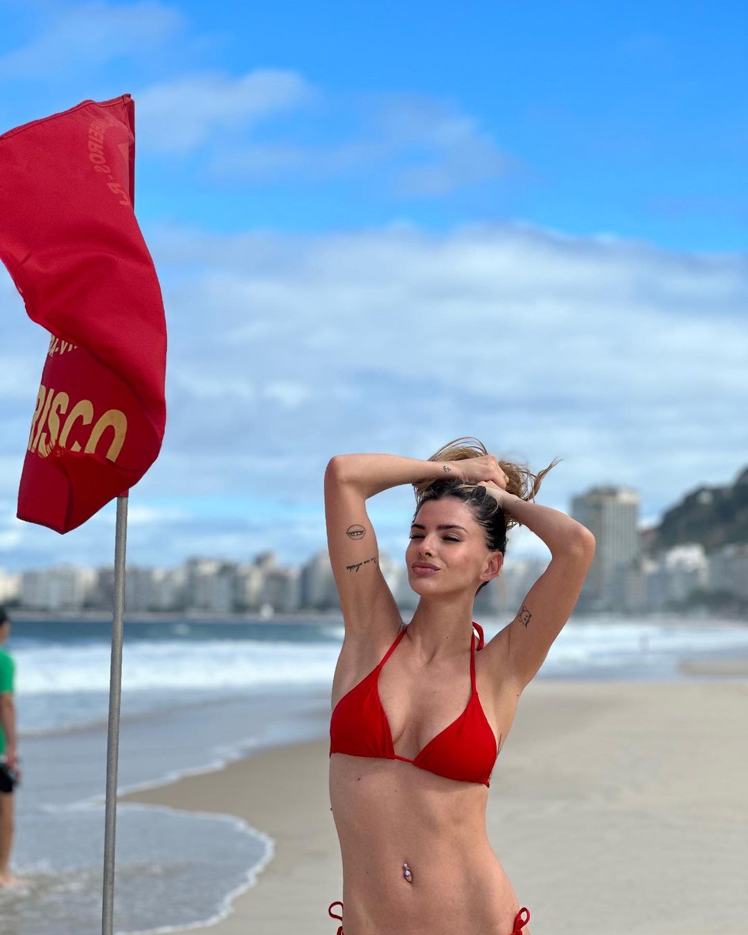 la china suarez con bikini rojo y las manos en la cabeza en una playa, una bandera roja a su lado / 