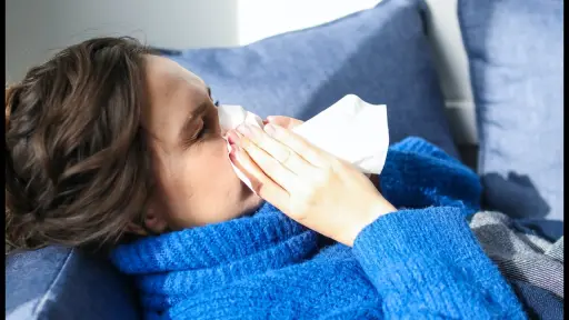 Una mujer estornuda mientras está sentada en un sofá debido a problemas de salud o falta de vitamina C