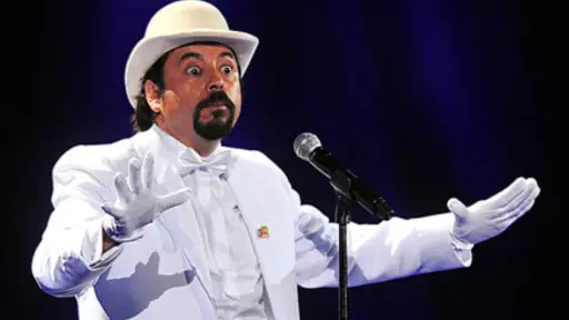 el humorista Bombo Fica de traje blanco y sombrero frente a un micrófono.