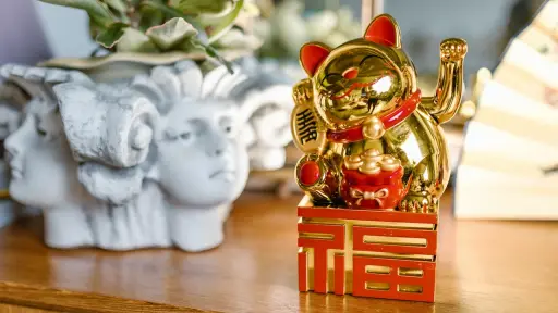 Un gato dorado de la suerte de cerámica sentado sobre una mesa de madera con un fondo borroso, como representación del elemento Feng Shui de prosperidad y abundancia para atraer la buena fortuna al hogar