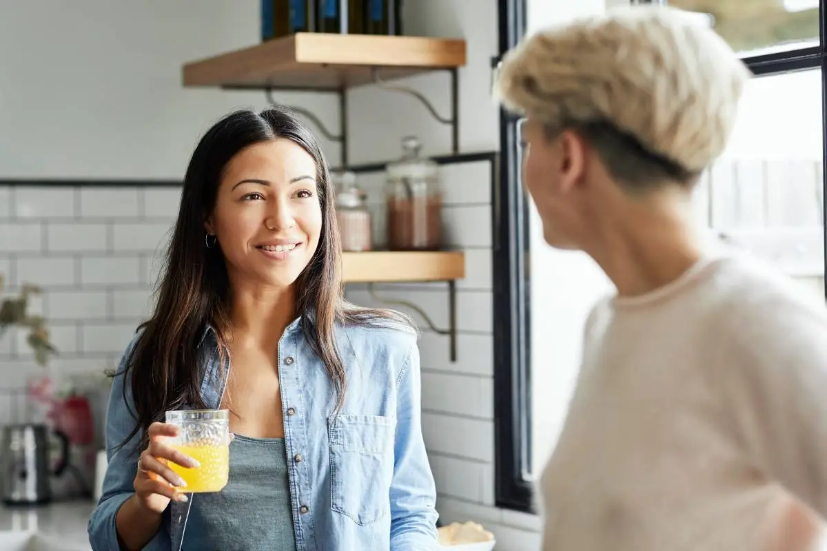 Woman Talking with Girlfriend While Holding Juice, Smiling young woman talking with girlfriend while holding juice. couple is spending leisure time at home. They are wearing casuals.