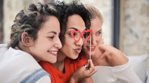 tres mujeres de 30 años posando para una fotografía y la del centro sosriene unos anteojos con forma de corazón