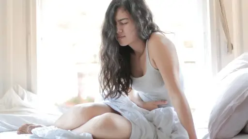 una mujer sentada en la cama se toma la barriga en señal de malestar como ejemplo de Síndrome de vómitos cíclicos