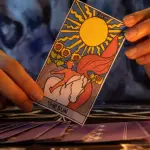 manos sosteniendo una carta del tarot sobre una mesa con velas y un mazo